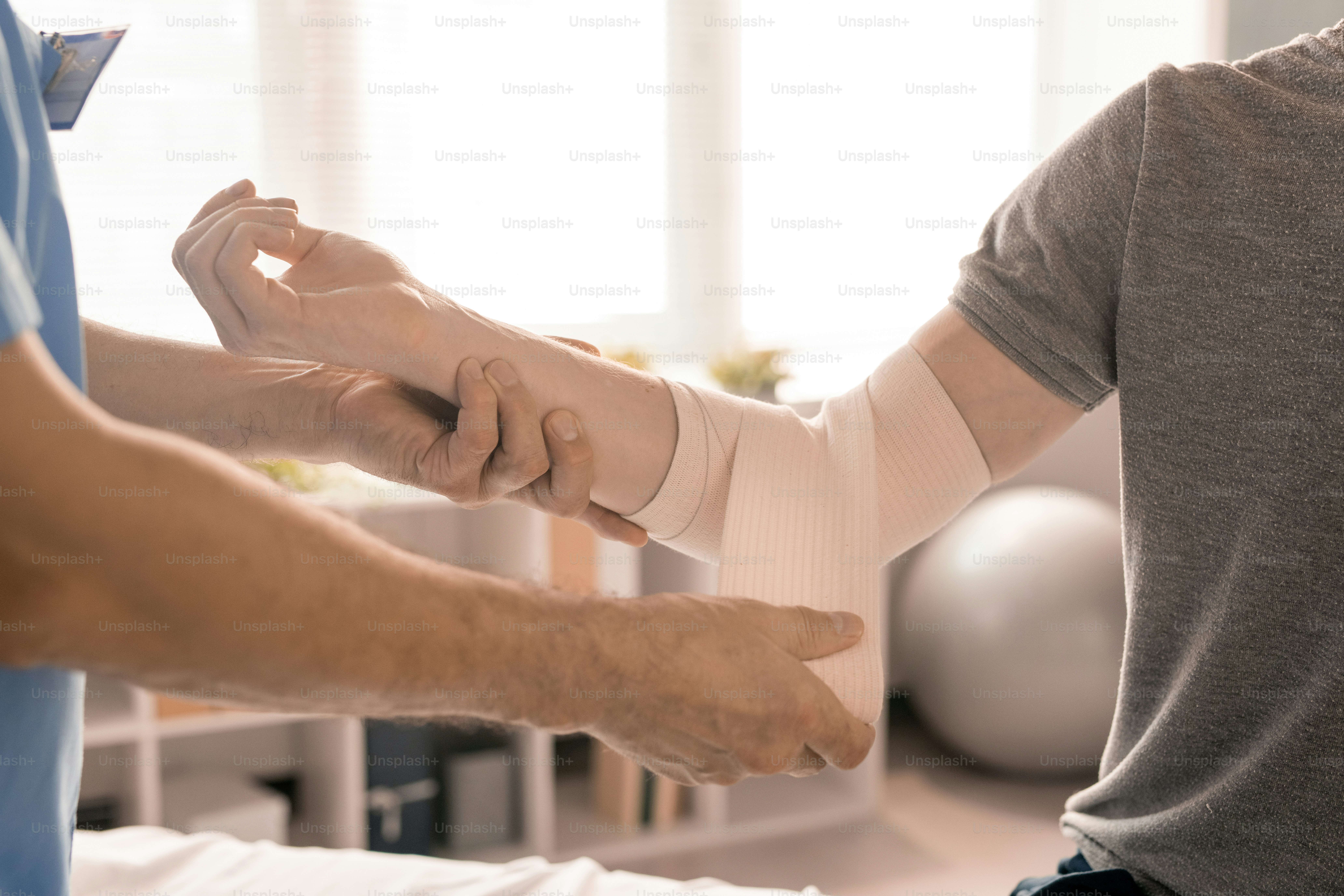 Hands of male clinician wrapping right elbow of sick young patient with flexible bandage during medical procedure in modern clinics